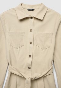 Beigefarbenes Cordkleid mit Kragen, langen Ärmeln, durchgehender Knopfleiste und zwei Brusttaschen. Enthält einen passenden Stoffgürtel.