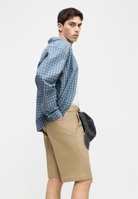 Νεαρός άνδρας με μπλε καρό πουκάμισο πάνω από λευκό τοπ και μπεζ σορτς, κρατώντας μαύρο καπέλο μπέιζμπολ στο πλάι, κοιτάζοντας πάνω από τον ώμο.