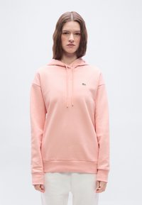 Ženska z rjavimi lasmi, oblečena v svetlo roza kapucar s majhnim zelenim logotipom krokodila in belimi hlačami na belem ozadju.