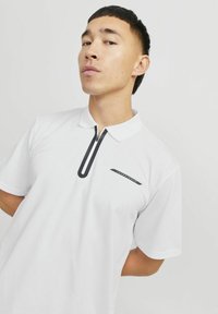 Hvid polo skjorte med en sort lynlås og accent langs forsiden. Har et diskret logo design på brystet og korte ærmer.