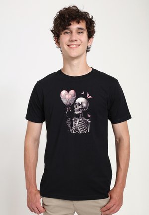 Smilende ung mand iført sort t-shirt med et skelet, der holder en lyserød hjerteformet ballon, omgivet af sommerfugle.
