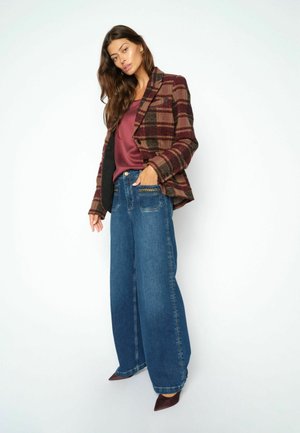 Vrouw met een geruite bruine jas, een bordeauxrode blouse, blauwe wijde spijkerbroek met kettinkdetail en puntige bruine schoenen, staand tegen een witte achtergrond.
