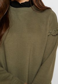 Mujer vistiendo un suéter verde oliva con cuello de encaje negro y detalles de encaje en la parte superior del brazo, con el cabello largo y marrón visible en el lado izquierdo.