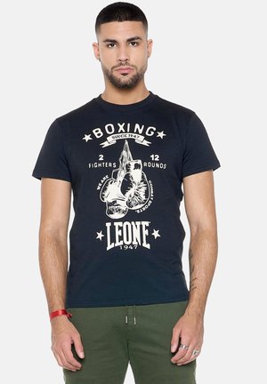 Uomo con capelli corti e barba che indossa una maglietta nera a tema pugilato e pantaloni verdi, in piedi davanti a uno sfondo chiaro uniforme.