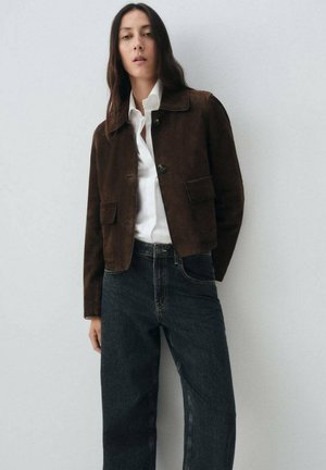 Chaqueta corta de ante marrón con dos bolsillos frontales, cierre de botones y cuello con solapa; llevada sobre una camisa blanca y jeans oscuros.