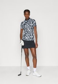 Polo de golfe com padrão de zebra em preto e branco, com gola e mangas curtas, combinado com uma saia preta. O modelo segura um taco de golfe e usa sapatos brancos.