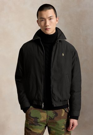 Jeune homme portant une veste noire zippée et un pull noir à col roulé, avec un pantalon camouflage vert et marron, debout devant un fond neutre.