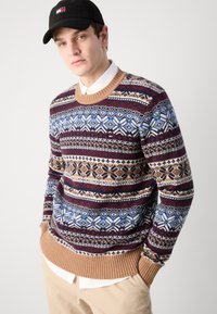 Braun, rot und blau gemusterter Strickpullover mit geometrischen Motiven und gerippten Bündchen, über einem weißen Hemd mit Kragen getragen.