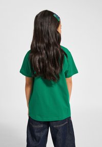 Grön bomullströja med korta ärmar, rak skärning och mjuk textur, i kombination med mörka jeans; långt vågigt hår synligt.