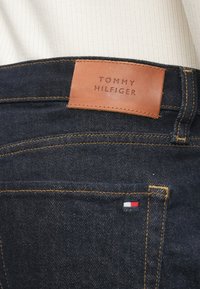 Vaqueros de mezclilla azul oscuro con una etiqueta de cuero marrón que presenta "TOMMY HILFIGER." Costuras naranjas notables y un pequeño detalle de bandera multicolor en el bolsillo.