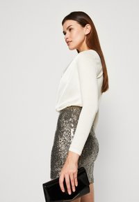 Weiße langärmelige Bluse, kombiniert mit einem metallischen Pailletten-Minirock. Das Outfit wird durch eine schwarze Clutch-Tasche ergänzt, was einen strukturellen Kontrast schafft.