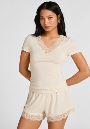 Femme aux longs cheveux foncés portant un haut beige à manches courtes et un short avec bordure en dentelle, debout devant un fond clair uni.