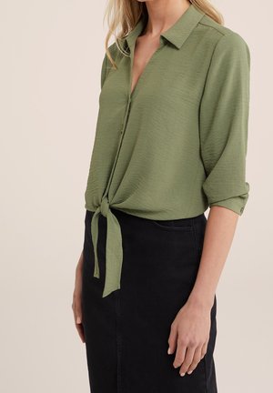 Femme portant un chemisier vert boutonné noué à la taille avec des manches retroussées et une jupe noire taille haute, debout sur un fond clair.
