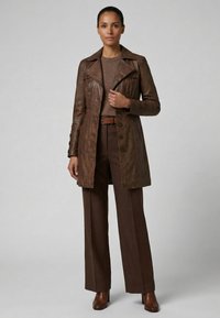 Kvinde stående iført en brun læderjakke, brun sweater, brede brune bukser, bælte og brune ankelstøvler mod en ensfarvet baggrund.