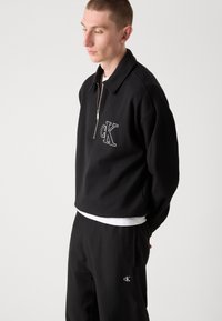 Crni zip-up dukserica s širokim okovratnikom i bijelim "CK" logom sprijeda, u kombinaciji s crnim hlačama koje imaju mali detalj s logom.