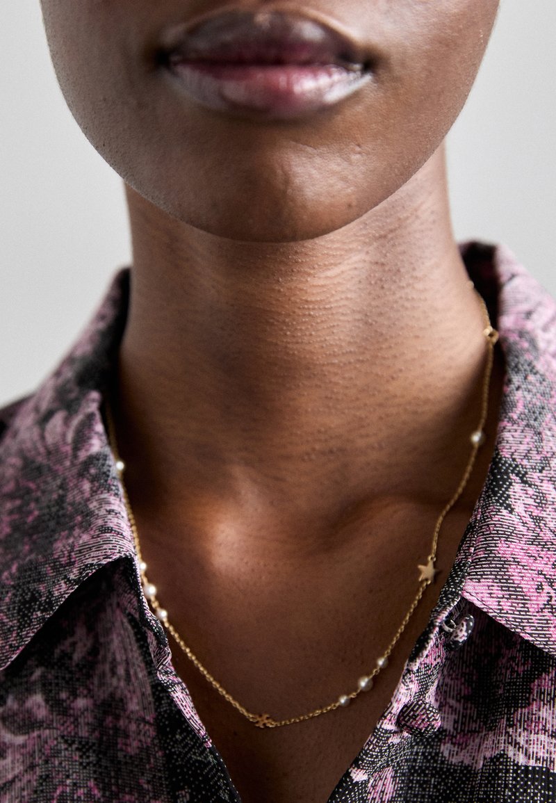 Zlata verižica z zvezdnimi detajli in belimi biseri, nošena čez srajco s patternom in ovratnikom. Posnetek v bližini območja vratu.