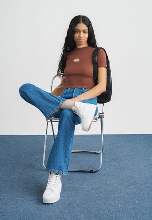 Rjava prilagojena majica z logotipom, modre razširjene kavbojke, bele superge in nogavice z vzorcem. Sedi na kovinskem stolu pred enostavnim ozadjem.