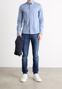 Uomo con una camicia azzurra chiara a bottoni, jeans blu, sneakers bianche, che tiene in mano a sinistra un giubbotto scuro piegato, in piedi su un pavimento chiaro.