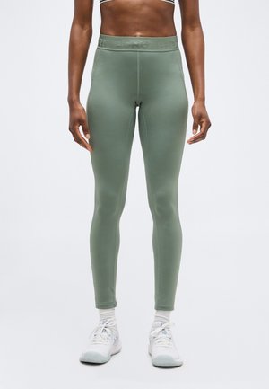 Κολάν σε στενή εφαρμογή σε ελιά, φορεμένα από έναν όρθιο άνθρωπο, σε συνδυασμό με λευκά αθλητικά παπούτσια και κάλτσες, πάνω σε απλό φόντο.