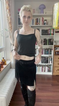 Top preto sem mangas e uma saia mini de veludo preta. A modelo usa botas pretas até ao joelho. O fundo apresenta estantes de livros e itens decorativos.