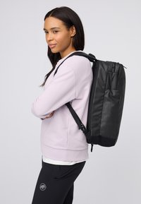 Kvinna i ljuslila sweatshirt och svarta byxor med en svart ryggsäck, står med armarna i kors mot en neutral bakgrund.