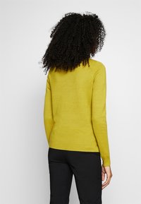 Personne aux cheveux bouclés portant un pull à manches longues jaune moutarde et un pantalon noir, debout de dos sur un fond uni.