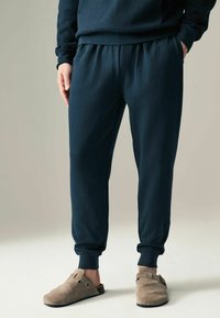 Marineblauwe sweatpants van zachte stof, met een elastische tailleband, boorden en zijzakken, gecombineerd met beige suède instapkleding.
