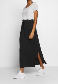 Jupe longue noire avec fentes latérales, associée à un haut à manches courtes rayé blanc et bleu marine, portée avec des baskets blanches de sport.