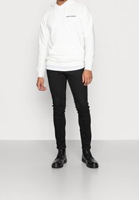 Vitt hoodie med en framficka och svart text, kombinerat med svarta slim-fit jeans och svarta snörskor, stående mot en neutralt bakgrund.
