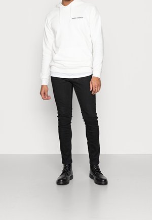 Sudadera blanca con un bolsillo frontal y texto negro, combinada con jeans ajustados negros y botas de cordones negras, de pie sobre un fondo simple.