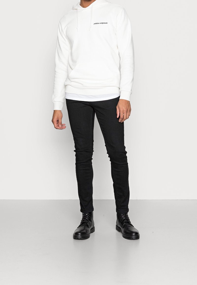 Vitt hoodie med en framficka och svart text, kombinerat med svarta slim-fit jeans och svarta snörskor, stående mot en neutralt bakgrund.