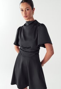 Crna haljina kratkih rukava s visokim ovratnikom i nabranim strukom, koja ima lepršavu suknju i glatku, sjajnu tkaninu. Jednostavan dizajn bez uzoraka.