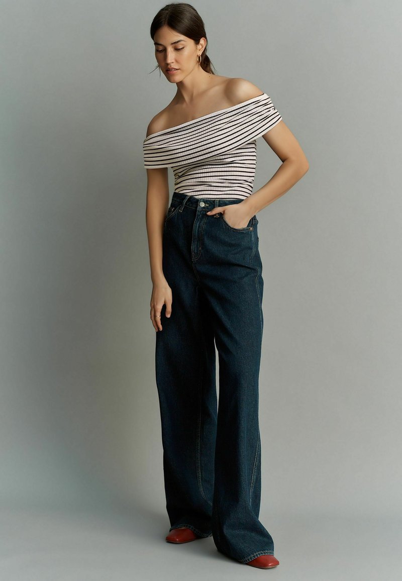 Gestreepte off-the-shoulder top in crème en zwart, gecombineerd met hoge taille donkerblauwe wijde jeans. Bruine schoenen maken de look compleet.