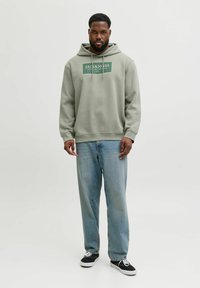 Mężczyzna z brodą w oliwkowej bluzie Jack & Jones, jasnoniebieskich dżinsach i czarnych sneakersach stoi na tle jasnego, jednolitego tła.