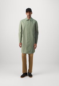 Impermeabile verde chiaro, lungo fino al ginocchio, con un design minimale, abbinato a pantaloni beige e sneakers nere. Presenta un colletto e chiusura a bottoni.