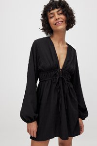 Čierne šaty s textúrovaným vzorom, hlbokým výstrihom do V a pásom na sťahovanie. Dlhé volánové rukávy so zahnutými manžetami. Dĺžka nad kolená.