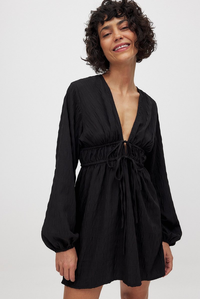 Čierne šaty s textúrovaným vzorom, hlbokým výstrihom do V a pásom na sťahovanie. Dlhé volánové rukávy so zahnutými manžetami. Dĺžka nad kolená.