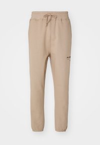 Beige szabású joggerek, elasztikus derékpánttal, húzózsinórral, enyhén beszűkülő lábakkal és fekete logóval a bal combon. Puha szövettextúra.