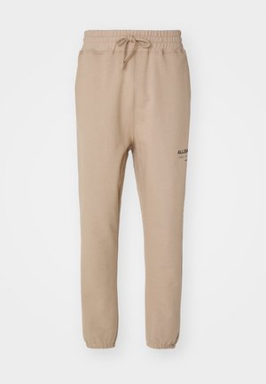 Beige Jogginghose mit elastischem Bund, Kordelzug, schmal zulaufenden Beinen und schwarzem Logo-Detailing am linken Oberschenkel. Weicher Stoff.