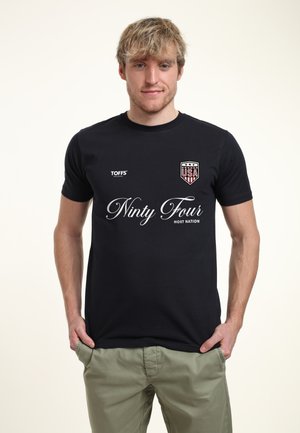 Giovane uomo con capelli biondi che indossa una t-shirt nera con emblemma USA e la scritta "Ninty Four", in piedi con le mani in tasca davanti a uno sfondo semplice.