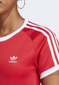 Czerwona koszulka sportowa Adidas z białymi, charakterystycznymi trzema paskami na ramionach i żebrowanym kołnierzykiem, z małym białym logo na klatce piersiowej.