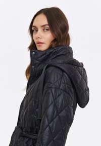Czarna, pikowana kurtka puchowa z kapturem, wyposażona w pasek nawiązujący, zapięcia na zatrzaski oraz błyszczącą teksturę, przedstawiona z boku na białym tle.
