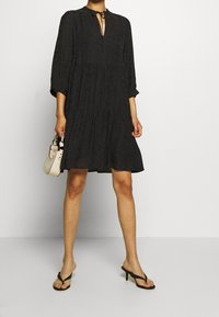 Vestido negro con diseño de volantes, textura plisada, mangas largas, un lazo en el cuello y un corte relajado. Lo lleva con chanclas negras y sostiene un pequeño bolso beige.