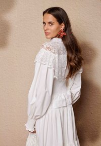 Λευκό φόρεμα με φουσκωτά μανίκια και δαντέλα στους ώμους· φούστα και μέση με βολάν. Μεγάλα κόκκινα σκουλαρίκια με λουλουδένιο μοτίβο προσθέτουν αντίθεση.