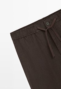 Pantalon marron foncé à taille élastique avec bouton à l'avant, cordon de serrage et tissu texturé, présenté de la taille jusqu'en bas.