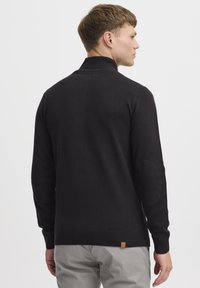 Pull noir en tricot avec un col montant, des poignets et un ourlet côtelés, comportant des renforts aux coudes et une petite étiquette de marque à la taille.