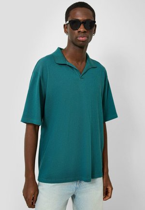 Jeune homme portant des lunettes de soleil noires, un polo à manches courtes bleu sarcelle et un jean bleu clair, debout devant un fond blanc uni.