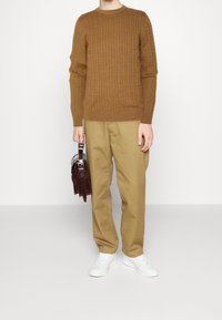 Texturiertes braunes Strickpullover mit gerippten Bündchen, kombiniert mit beigen Hosen. Weiße Sneaker und eine kleine dunkelbraune Tasche vervollständigen das Outfit.