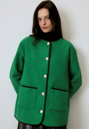 Kvinde med langt mørkt hår iført en struktureret grøn knappet jakke med sort kant og to forlommer, stående mod en ensartet hvid baggrund.