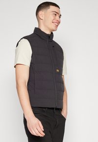 Svart vadderad väst med hög krage, framsidan dragkedja och liten logodetalj. Bärs över en ljus t-shirt, i kombination med svarta byxor.